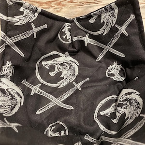 May include: Black bowl cozy with a white pattern of swords, wolf heads, and other symbols. The cozy is quilted and has a square shape with a folded-over edge. The design is repeated across the fabric.