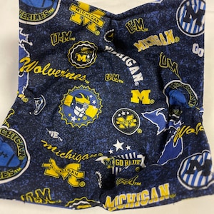 May include: A navy blue and yellow bowl cozy with a pattern of the University of Michigan Wolverines logo and text. The cozy is square-shaped and made of fabric. The text includes "MICHIGAN", "Wolverines", and "Go Blue".