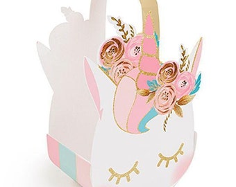 May include: A white paper gift basket with a handle, featuring a unicorn face design. The unicorn has closed eyes with gold eyelashes, a pink and gold horn, and a floral arrangement in shades of pink, gold, and teal on its forehead.