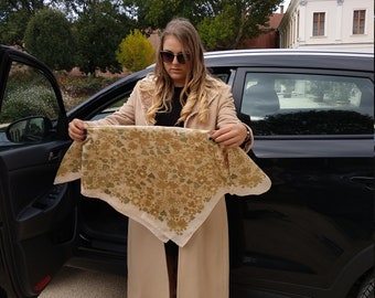 May include: A woman wearing a beige coat and sunglasses holds a beige and brown floral patterned scarf in front of a black car.