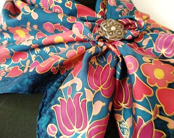 Puede incluir: Un pañuelo de seda azul turquesa con un estampado floral rosa y dorado. El pañuelo está anudado y tiene un broche de plata con un diseño floral.