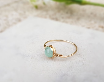 May include: A delicate gold ring featuring a light green, faceted gemstone. The gemstone is wire-wrapped to the gold band. The ring is displayed on a white surface, with a soft-focus background.