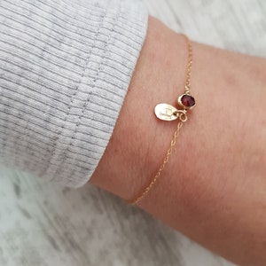 Puede incluir: Un brazalete de cadena dorada con una pequeña piedra preciosa roja y un dije con la letra "L" grabada.