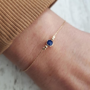 Könnte beinhalten: Ein zartes Armband aus einer goldenen Kette mit einem einzigen blauen Edelstein. Der Edelstein ist facettiert und hat eine runde Form.