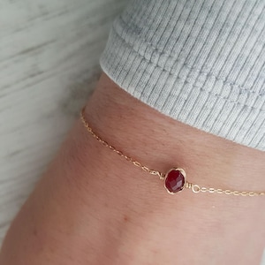Puede incluir: Un delicado brazalete de cadena dorada con una sola piedra preciosa roja. La piedra preciosa es de forma ovalada y está engastada en un simple alambre de oro.