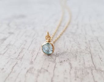 May include: A delicate gold necklace featuring a light blue gemstone pendant. The pendant is a raw, faceted stone with a gold setting.