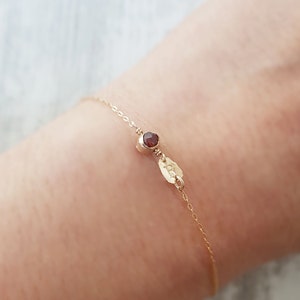 Puede incluir: Un delicado brazalete de cadena dorada con una pequeña piedra preciosa roja y un dije de oro. El brazalete está en la muñeca de una persona.