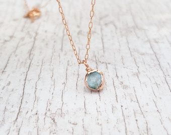 May include: A delicate gold chain necklace featuring a small, light blue gemstone pendant. The gemstone is wrapped in gold wire and has a natural, irregular shape.