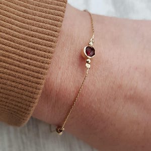 Könnte beinhalten: Ein zartes Armband aus einer goldenen Kette mit einem einzigen roten Edelstein. Der Edelstein ist in einer goldenen Fassung gefasst und von kleineren goldenen Perlen umgeben.