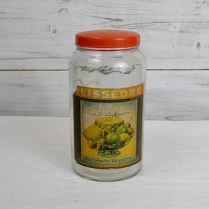 May include: A clear glass jar with an orange lid. The jar has a label with the text "TEISSEDRE" and a picture of fruit. The label also includes the text "à la Liqueur" and "Etab. André Teissèdre - BORDEAUX".