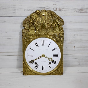 Puede incluir: Reloj de pared antiguo de color dorado con esfera blanca y números romanos negros. El reloj presenta una talla en relieve ornamentada y detallada de figuras y follaje en la parte superior. La esfera del reloj dice "Sylvain BACHET" y "J. Lemaire".