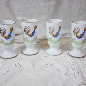 Peut inclure: Ensemble de quatre tasses en céramique blanche avec un motif de coq. Chaque tasse présente un coq bleu avec des accents jaunes et une queue brune, debout sur une base verte. Les tasses ont un bord brun et une base blanche.