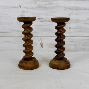 Puede incluir: Dos candeleros antiguos de madera. Cada soporte tiene un tallo torneado en espiral y una base redonda. Los candeleros están hechos de madera de color marrón oscuro y se exhiben sobre una superficie blanca.