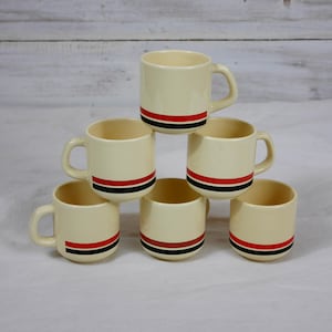 Peut inclure: Six tasses en céramique crème empilées. Chaque tasse a une anse et une bande rouge et noire au milieu. Idéales pour le café ou le thé, elles sont disposées en pyramide.
