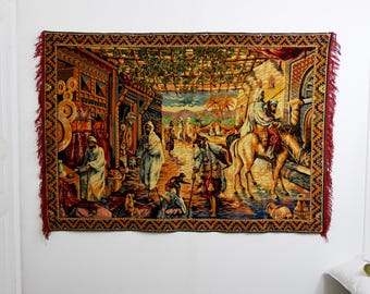 May include: A rectangular tapestry depicting a Middle Eastern marketplace scene. The tapestry features figures in traditional attire, buildings, and palm trees. The color palette includes reds, browns, and yellows, with a fringed border.