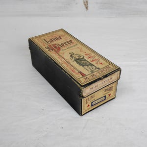 Puede incluir: Una caja negra rectangular vintage con una etiqueta decorativa. La etiqueta presenta el texto "Laine S Pierre" y una ilustración de un hombre. La caja parece ser de cartón.