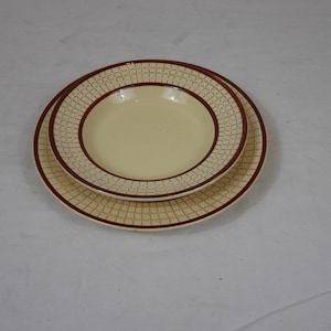 Peut inclure: Ensemble de vaisselle beige et marron en deux pièces. L'ensemble comprend une assiette et un petit bol. Les deux pièces ont un centre beige et un bord marron avec un motif carré répétitif. Les assiettes sont empilées.