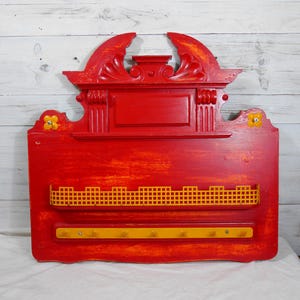 Puede incluir: Estante de pared de madera roja con una estantería de rejilla amarilla y ganchos amarillos. La estantería tiene una parte superior decorativa con un diseño tallado.