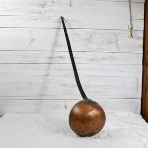 May include: A vintage copper and iron cooking pot with a long, curved handle. The pot has a round, bulbous copper base and a dark iron handle. The handle extends upwards and curves gracefully, ending in a small loop. The pot is likely used for cooking.