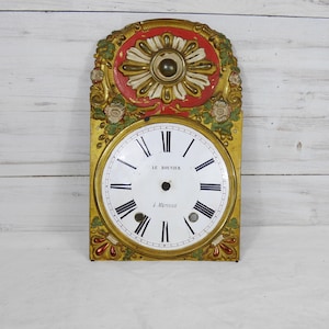 Puede incluir: Esfera de reloj antigua con esfera blanca, números romanos negros y el texto "LE BOUVIER à Mariannet". La esfera del reloj está enmarcada en dorado, rojo y verde con detalles florales y ornamentados. La esfera del reloj es rectangular.