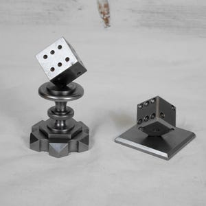 Könnte beinhalten: Zwei Würfelskulpturen aus Metall. Ein Würfel ist auf einem Sockel mit geometrischer Basis und zylindrischem Schaft montiert. Der andere Würfel ruht auf einer quadratischen Basis. Beide Würfel haben sichtbare Punktmuster.