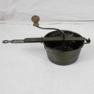 Puede incluir: Un molinillo de alimentos vintage de metal con un mango de madera. El molinillo tiene un cuenco redondo perforado y una manivela con una perilla de madera. El molinillo se utiliza para colar y hacer puré los alimentos.