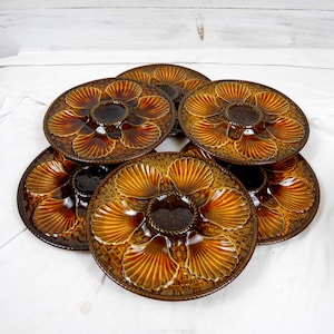 May include: A collection of six brown ceramic oyster plates. Each plate features a central well and six shell-shaped compartments. The plates have a textured, woven-like pattern and are arranged on a white surface.