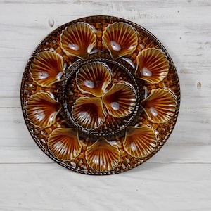 May include: A brown ceramic oyster serving platter with shell-shaped compartments. The platter has a woven texture around the edges and a central section with smaller shell-shaped dishes. The shells are a warm brown color.