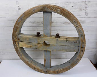 May include: A large, wooden wheel with a center hole and four spokes. The wheel is painted a light blue color and has a natural wood finish on the rim. The wheel is weathered and shows signs of age.