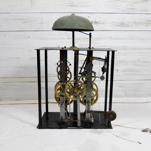 Puede incluir: Un antiguo mecanismo de reloj con engranajes de latón visibles y una campana verde. El marco de metal negro alberga los intrincados mecanismos del reloj. El mecanismo del reloj se encuentra sobre un fondo de madera blanca.