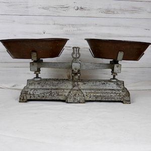 Peut inclure: Balance ancienne en métal blanc avec deux bols en cuivre. La balance a un aspect usé et rustique, parfait pour une cuisine vintage ou une décoration de ferme.