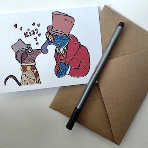 May include: A greeting card with a cartoon illustration of two characters kissing, with the word "Kiss" and heart shapes above them. The card is accompanied by a brown envelope and a silver pen.