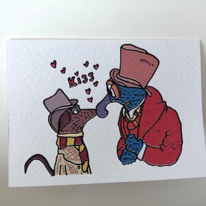 May include: A greeting card featuring two cartoon characters kissing. One is a blue creature in a red coat and top hat, the other is a brown dog wearing a scarf and hat. The word "Kiss" is above them with hearts.