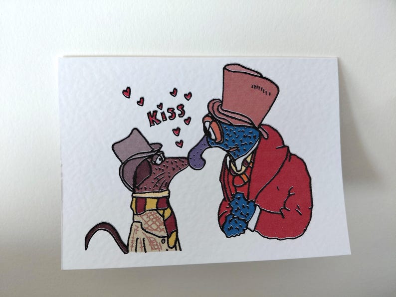 May include: A greeting card featuring two cartoon characters kissing. One is a blue creature in a red coat and top hat, the other is a brown dog wearing a scarf and hat. The word "Kiss" is above them with hearts.
