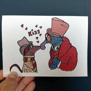 May include: A whimsical greeting card featuring two cartoon characters about to kiss. One is a dog wearing a hat and scarf, the other a blue-skinned creature in a red coat and top hat. The word "Kiss" is written above them with hearts.