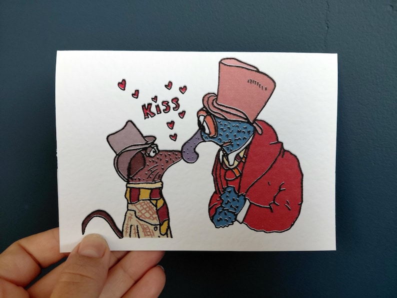 May include: A whimsical greeting card featuring two cartoon characters about to kiss. One is a dog wearing a hat and scarf, the other a blue-skinned creature in a red coat and top hat. The word "Kiss" is written above them with hearts.