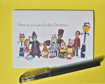 May include: A white greeting card with the text "Have an uncontrollable Christmas." features a whimsical illustration of costumed characters and street lamps. A black pen with a clear cap rests on a yellow surface.