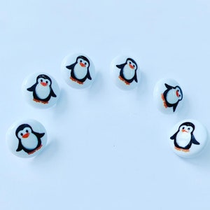 May include: White ceramic buttons with a hand-painted penguin design. Each button features a black penguin with orange feet and beak, and a white belly. The buttons are arranged on a white surface.