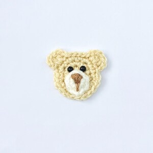 May include: A cream-coloured crocheted bear face applique with brown and black details. The bear features small black eyes, a brown nose, and a cream-coloured muzzle. The ears are rounded, creating a simple and charming design.