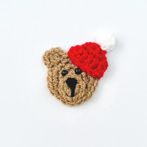 May include: A crocheted teddy bear face wearing a red and white Santa hat. The bear is a light brown colour with black eyes and nose. The hat is red with a white bobble. The background is white.