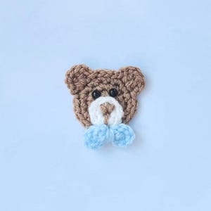 May include: A crocheted teddy bear applique. The bear is brown with a white muzzle and a light blue bow tie. The bear has black button eyes. The background is a light blue.