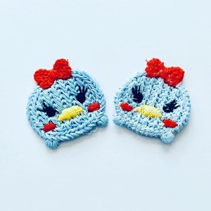 May include: Two blue embroidered patches with red bows, yellow beaks, and black eyes. The patches are shaped like a cartoon bird.