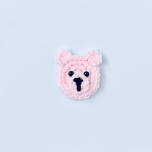 May include: A pink crocheted bear face with black eyes and a black nose.