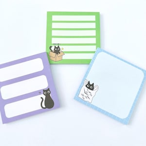 May include: Three square sticky note pads in green, purple, and light blue. The green pad has white horizontal lines and a black cat in a box with the text KISS ME. The purple pad has three white rectangular areas and a black cat. The blue pad has a grid border and a black cat in a bag.