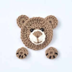 May include: Crocheted brown bear applique with a white face and black eyes. The bear has two small brown crocheted paws with black claws.
