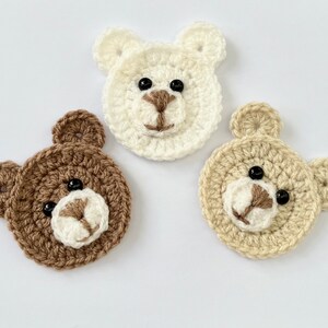 May include: Three crocheted bear faces in brown, beige, and white. Each face has black eyes and a white nose.