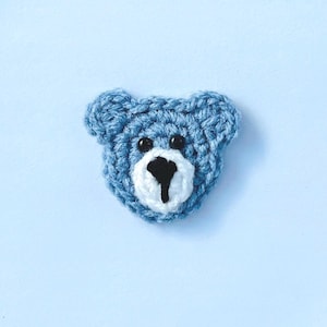 May include: A crocheted blue teddy bear face with black eyes and a white and black muzzle. The bear is set against a light blue background. The handmade bear is a charming craft item.