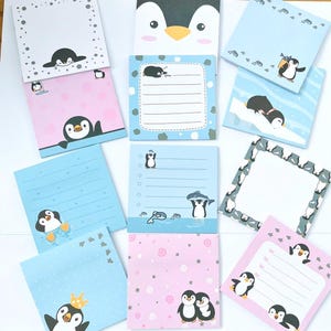 May include: A collection of square sticky note pads featuring various penguin illustrations. The designs include penguins in blue, pink, and white colour schemes, some with lined writing areas, others with decorative borders, and cartoon motifs of penguins swimming, fishing, or wearing crowns.