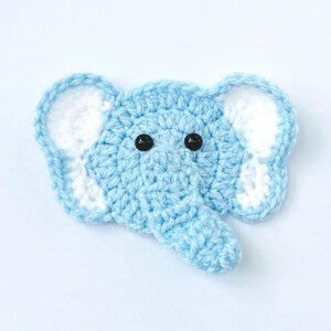May include: A light blue crocheted elephant head with white ear detailing. The elephant has black button eyes and a trunk. This handmade item is suitable for baby showers, nurseries, or craft projects.