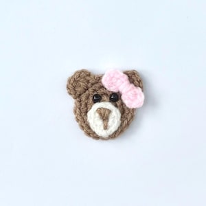 May include: A crocheted bear face with a pink bow. The bear is brown with a white muzzle and black eyes. The bow is a light pink colour. The bear is a handmade item.